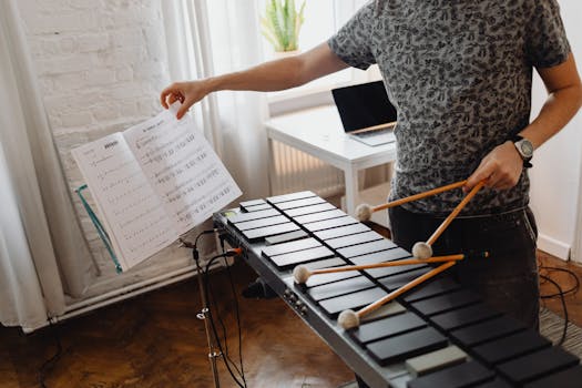 A person plays a xylophone while turning pages of a music book indoors.